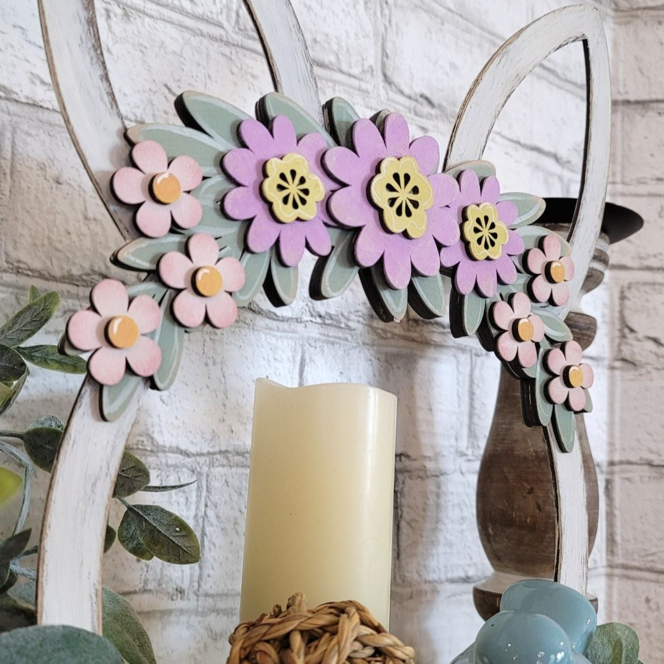 Charming wooden Easter tray with distressed chalk paint finish, featuring purple and pink flowers on a green leaf swag, and Easter bunny ears. Hand-painted and laser cut, this 2-sided tray can be enjoyed from all angles and is perfect for displaying candles with flowers or cookies. The tray also features unique repurposed sewing thread spools for feet, adding to its charm and eco-friendliness.