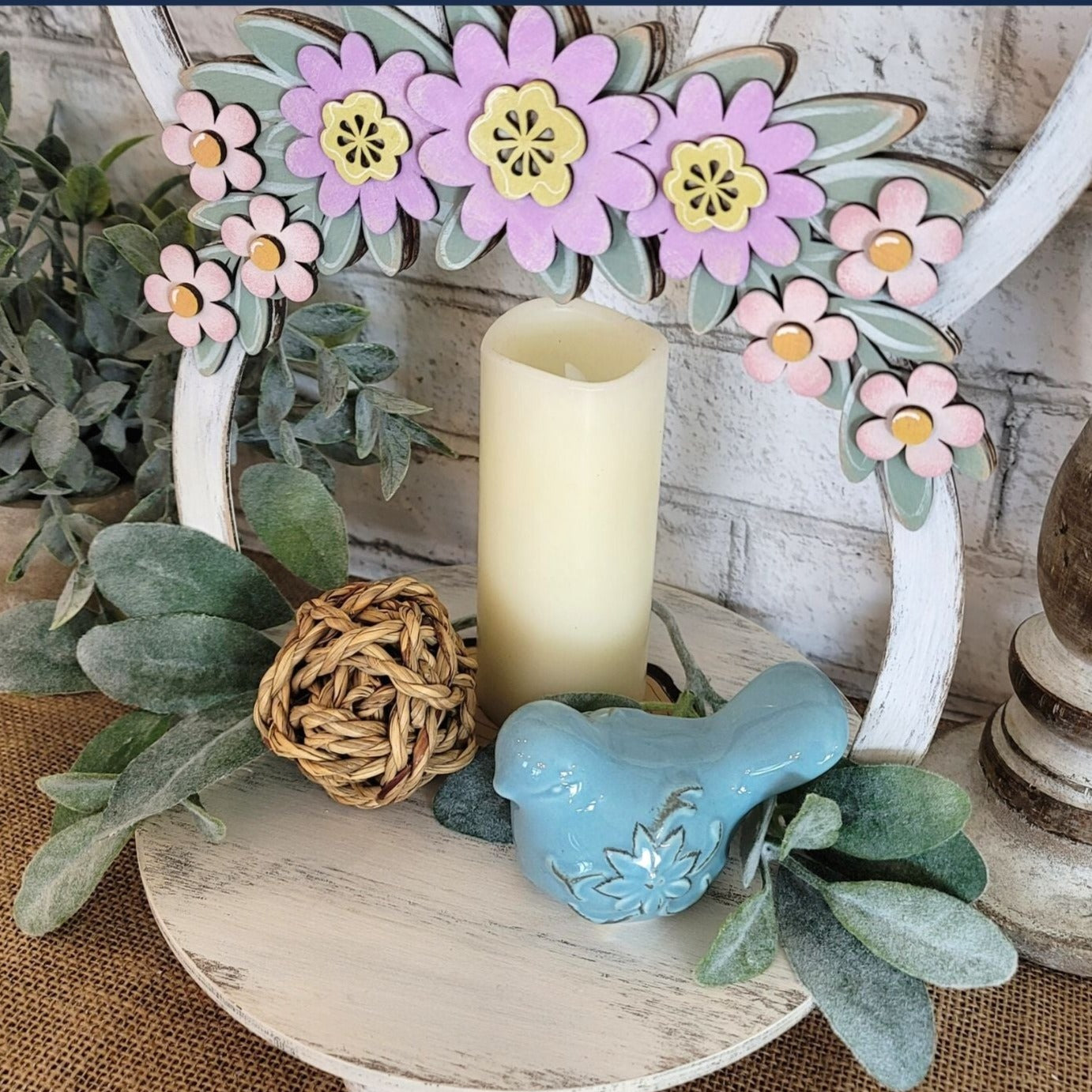 Charming wooden Easter tray with distressed chalk paint finish, featuring purple and pink flowers on a green leaf swag, and Easter bunny ears. Hand-painted and laser cut, this 2-sided tray can be enjoyed from all angles and is perfect for displaying candles with flowers or cookies. The tray also features unique repurposed sewing thread spools for feet, adding to its charm and eco-friendliness.