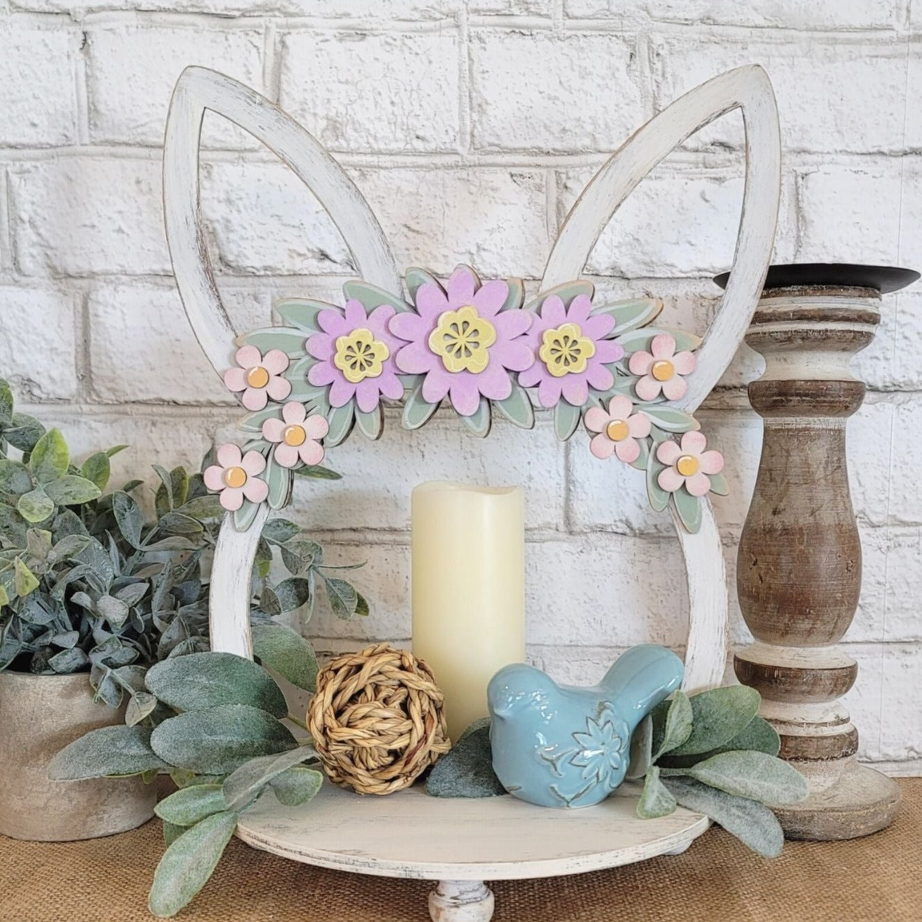 Charming wooden Easter tray with distressed chalk paint finish, featuring purple and pink flowers on a green leaf swag, and Easter bunny ears. Hand-painted and laser cut, this 2-sided tray can be enjoyed from all angles and is perfect for displaying candles with flowers or cookies. The tray also features unique repurposed sewing thread spools for feet, adding to its charm and eco-friendliness.