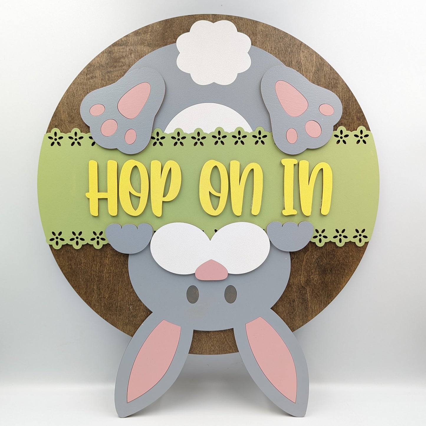15 inch hand-painted wooden round door sign featuring an upside down Easter Bunny and the message "Hop On In". Designed for display on a covered porch or entryway out of direct weather elements. Can be hung using attached jute twine or sawtooth hanger.