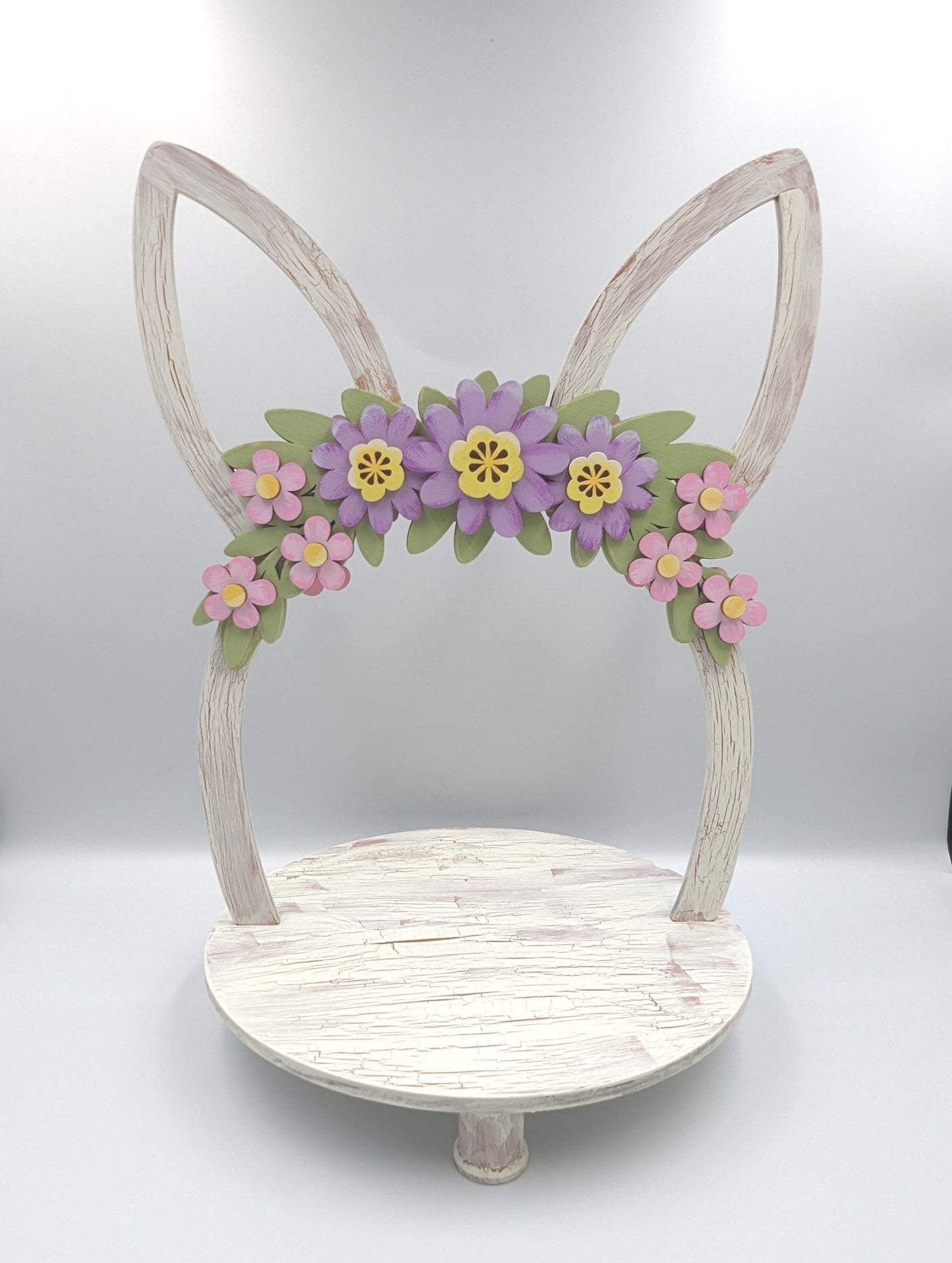Charming wooden Easter tray with distressed chalk paint finish, featuring purple and pink flowers on a green leaf swag, and Easter bunny ears. Hand-painted and laser cut, this 2-sided tray can be enjoyed from all angles and is perfect for displaying candles with flowers or cookies. The tray also features unique repurposed sewing thread spools for feet, adding to its charm and eco-friendliness.